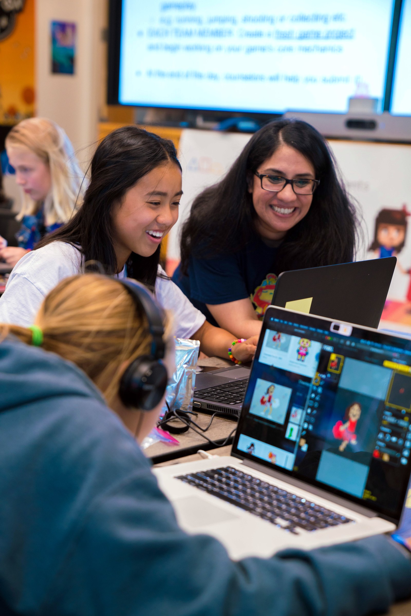 image shows a young woman smiling while the founder of Girls Make Games looks on over her left shoulder. Other girls are visible in the background and in the extreme foreground. They are working intently at laptops.