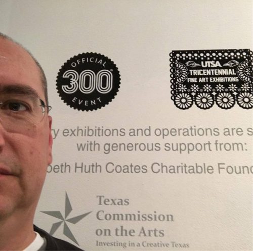 selfie of Rafael standing in front of UTSA Gallery wall texts that verify Deep Roots is part of the 300 anniversary celebrations of San Antonio, Texas